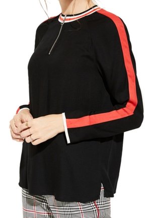 Femme portant un haut noir à manches longues avec des rayures rouges sur les manches, un col zippé et un pantalon à carreaux avec un motif noir, blanc et rouge.