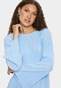 Blouse plissée bleu clair avec de longues manches amples et un col rond. Texture lisse avec des rayures verticales et un subtil éclat.