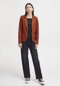 Roestkleurig, geruit blazer met een aansluitend ontwerp, gecombineerd met een zwarte top en losse zwarte broeken, gedragen met zwarte hoge sneakers.