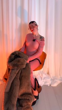 Roze geribbelde jurk met off-shoulder ontwerp, gecombineerd met een textuurrijke bruine bontsjaal. Zwarte open-toe schoenen met zigzagaccenten. Oranje achtergrond.