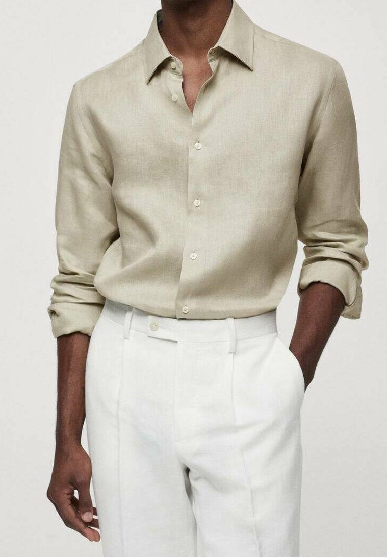 Personne à la peau foncée portant une chemise beige à boutons avec les manches retroussées et un pantalon blanc taille haute, une main dans la poche.