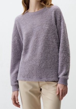 Femme portant un pull violet lavande à manches longues et duveteux avec un pantalon beige, debout devant un fond clair uni.
