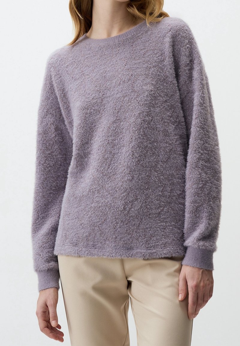 Femme portant un pull violet lavande à manches longues et duveteux avec un pantalon beige, debout devant un fond clair uni.