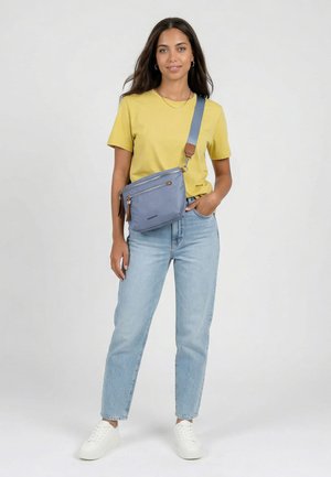 Kvinde iført gul t-shirt, lyseblå jeans, hvide sneakers og blå crossbody taske, står med den ene hånd i lommen og smiler til kameraet.