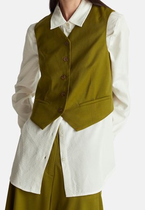 Femme portant un gilet boutonné vert olive par-dessus une chemise blanche texturée à manches longues, avec un pantalon assorti vert olive.