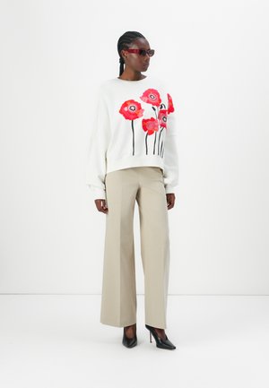 White sweater with red poppy embroidery, paired with beige wide-leg trousers and black high heels. Model wearing burgundy sunglasses.