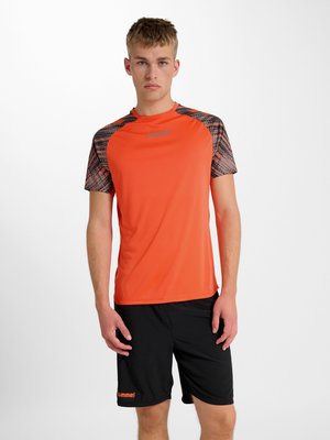 Ung mand stående iført en orange sportstrøje med mønstrede ærmer og sorte shorts med brandlogos på hvid baggrund.