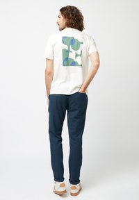 Witte katoenen T-shirt met groene en blauwe abstracte vormen op de achterkant. Gecombineerd met marineblauwe broek en lichtgekleurde sneakers.