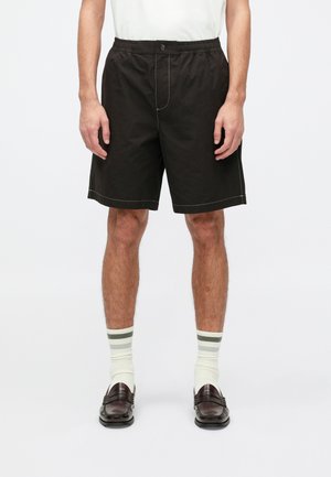 Pantaloni shorts neri con vestibilità rilassata, vita elastica e bottone frontale. Dettaglio di cucitura a contrasto. Indossati con calze a righe bianche e verdi.