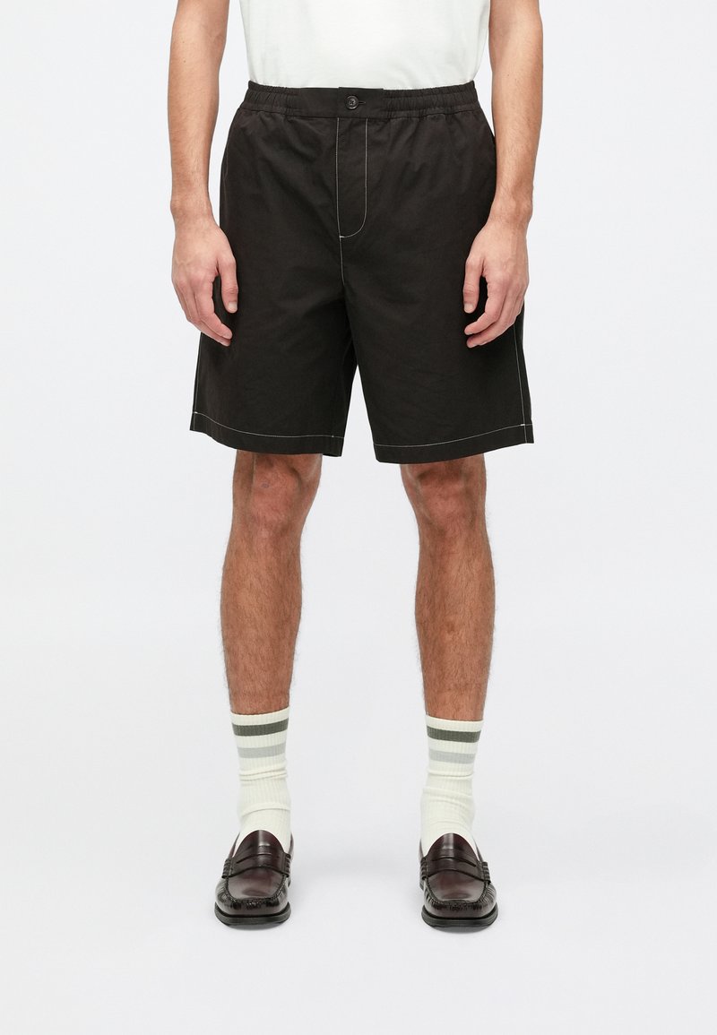 Shorts noirs à coupe décontractée, avec un élastique à la taille et un bouton à l'avant. Détail de couture contrastante. Portés avec des chaussettes rayées blanches et vertes.
