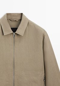 Beige afslappet jakke med krave, forrest lynlåslukning og lynlås-lomme på højre side, vist på hvid baggrund.