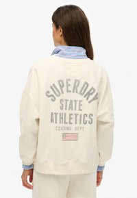 Crèmekleurige sweatstof met de tekst "Superdry State Athletics" in grijs, met een gestreepte blauwe en roze kraag die erbovenuit steekt.