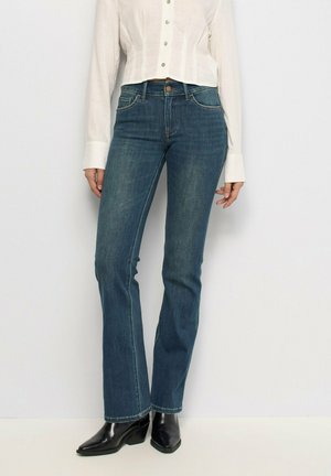 Mujer con vaqueros rectos azul oscuro, camisa blanca de manga larga con botones y botines negros de punta en un fondo blanco.