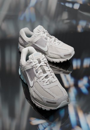 Par af hvide og grå Nike løbesko med mesh- og læderdetailler placeret på en reflekterende overflade med sløret baggrund.