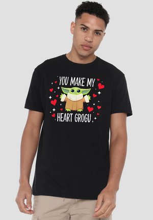 Junger Mann trägt ein schwarzes T-Shirt mit grünem Grogu-Charakter, umgeben von roten Herzen und dem Text „You make my heart Grogu“ vorne.