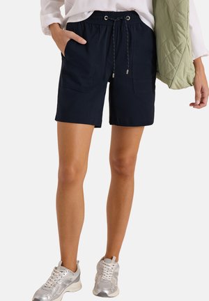 Kvinde iført navy shorts med snøre og forlommer, hvide sneakers, hvid langærmet top og holder en grøn quiltet jakke.