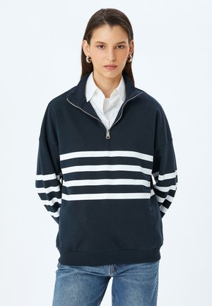 Marineblauwe sweatshirt met witte horizontale strepen, ritssluiting bij de kraag, losse pasvorm en geribde manchetten. Gekozen over een wit overhemd met kraag.
