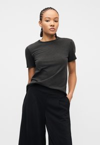 Νεαρή γυναίκα με πλεξούδες φοράει απλό σκούρο γκρι μπλουζάκι και φαρδιά μαύρα παντελόνια, στέκεται με το ένα χέρι στην τσέπη μπροστά σε λευκό φόντο.