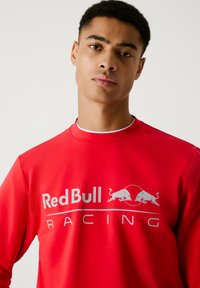 Młody mężczyzna z krótkimi włosami, ubrany w czerwoną bluzę z kapturem z białym logo Red Bull Racing na przodzie.