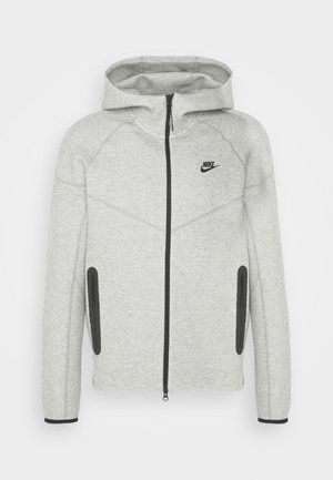 Felpa grigia con zip frontale, dettagli neri e cappuccio. Presenta maniche raglan e due tasche laterali con cerniera. Logo Nike visibile.