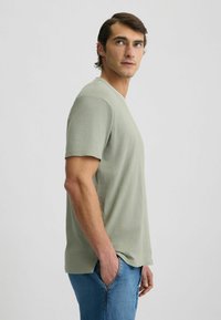 T-shirt verde chiaro a maniche corte con scollatura a girocollo, texture liscia e orlo dritto, indossato con jeans blu, mostrato da una vista laterale.