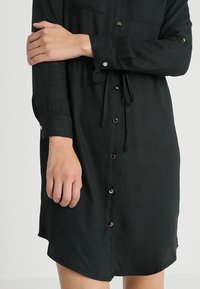 Tmavě zelené šaty z hladké tkaniny, vybavené knoflíkovými zapínáními, dvěma kapsami na hrudi a opaskem v pase s uvolněným střihem.