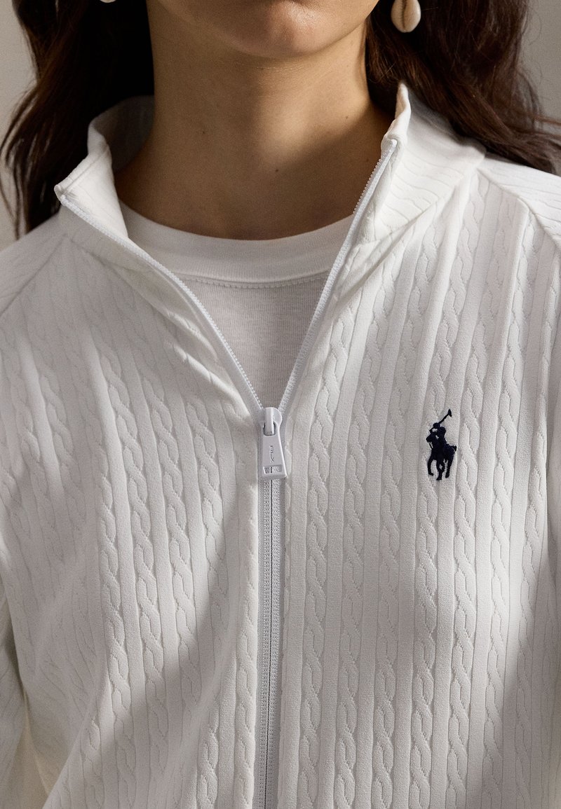 Femme portant un pull zippé en maille torsadée blanche avec un logo de joueur de polo brodé bleu marine sur la poitrine.