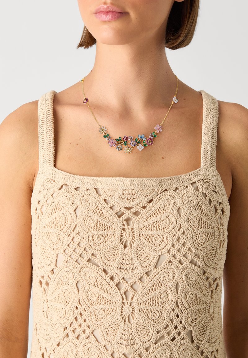 Femme portant un haut beige en crochet avec des motifs papillons et un collier chaîne en or orné de breloques florales multicolores.