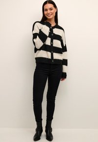 Svart och krämrandig kofta med en fluffig textur, med rund halsringning och framknappar, bärs tillsammans med svarta jeans och ankelstövlar.