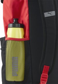 Zaino in tessuto nero, con accenti rossi e una tasca laterale in rete contenente una bottiglia d'acqua gialla. Presenta un'etichetta con logo sul manico.