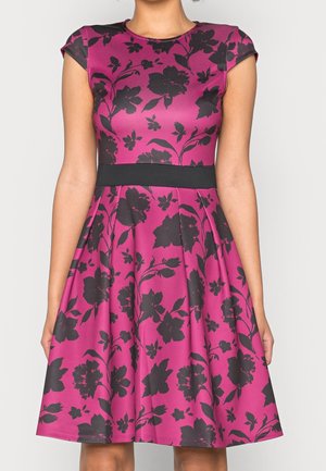 Kobieta ubrana w różową sukienkę do kolan z czarnym kwiatowym nadrukiem, z krótkimi rękawkami i czarnym paskiem w talii.