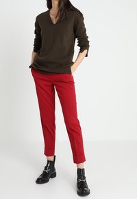 Donna che indossa un maglione a V marrone scuro, pantaloni rossi su misura alla caviglia e stivali neri alla caviglia, in piedi con le mani in tasca.