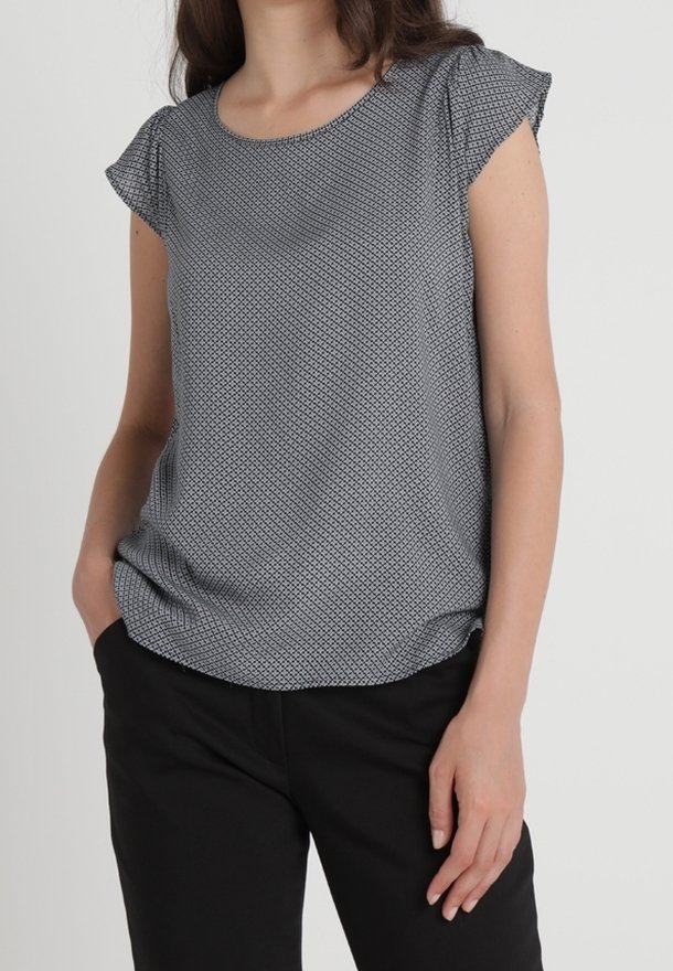 Femme portant un blouson gris à motifs à manches courtes avec un pantalon noir, se tenant avec une main dans la poche et l'autre détendue à ses côtés.