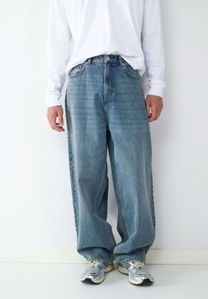 Persoon draagt losse blauwe spijkerbroek, wit shirt met lange mouwen en zilveren hardloopschoenen, staand tegen een witte muur.
