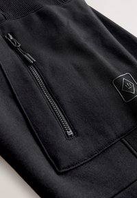 Zwarte sportieve joggers met een gladde stof, geribbelde tailleband, een zak met ritssluiting en een klein merkembleem van een contrasterend materiaal.