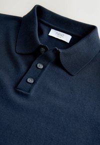 Tumesinine polosärk kraega, millel on kolm musta nööpi, sooniline tekstuur ja krae sees silt, millel on kiri „next”.