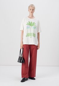 T-shirt blanche surdimensionnée avec un imprimé graphique vert et violet, associée à un pantalon large rouge et un sac à main noir. Texture de tissu lisse.