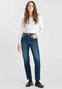 Jean bleu foncé avec une coupe droite, dotés d'une taille haute et d'un design à cinq poches. Associés à un haut blanc à manches longues et des chaussures noires.
