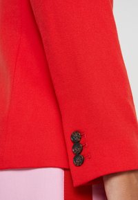 Punainen villaBleiserin hiha, jossa on kolme mustaa nappia ja sileä pinta, ja vaaleanpunainen kangas kurkistaa alhaalta.