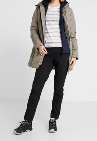 Parka gris clair avec capuche sur une couche intermédiaire zippée bleu marine, associée à une chemise blanche rayée et un pantalon noir ; chaussures de randonnée noires avec des accents turquoise.