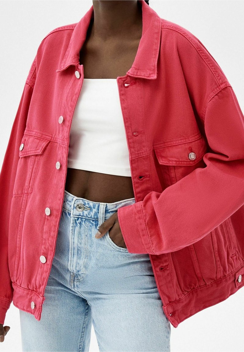 Chaqueta de denim rosa de gran tamaño con cierre de botones, dos bolsillos frontales y mangas largas. Combinada con unos pantalones vaqueros de tiro alto en azul claro.