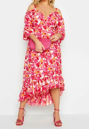 Kvinde iført en lyserød blomsterprint kjole med bare skuldre og flæset kant, der holder en tekstureret lyserød clutch og har matchende lyserøde hælede sandaler på.