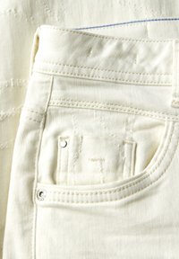 Vitt denimtyg med en närbild av en ficka, som har silverfärgade detaljer, synliga sömmar och subtila strukturerade mönster.