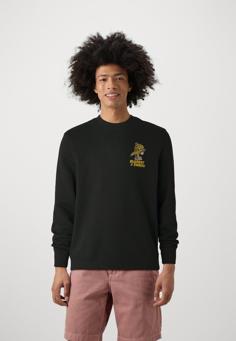 Zwart sweatshirt met lange mouwen, voorzien van een gele grafische print op de borst. De stof lijkt glad. Model draagt lichtroze shorts.