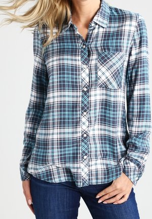 Femme portant une chemise à carreaux bleue, noire et blanche à boutons, manches longues, avec une poche poitrine unique, associée à un jean bleu foncé.