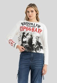 Weißer Langarm-Sweatshirt mit schwarzem und rotem Grafikdruck, featuring Motorradabbildungen und Markennamen. Getragen mit blauen Cargohosen.