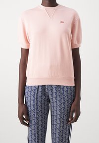 Jasnoróżowa bluza z krótkim rękawem z prążkowanym dekoltem i ściągaczem u dołu. Na piersi małe haftowane logo. Tkanina o fakturze, luźny krój.