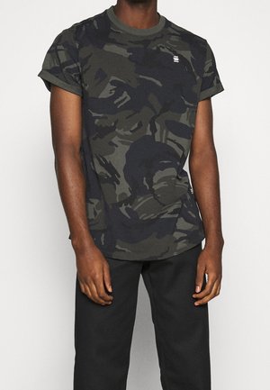 Mann trägt ein dunkelgrünes und schwarzes Camouflage-T-Shirt mit kurzen Ärmeln und schwarze Hose, steht vor einem einfarbigen Hintergrund.