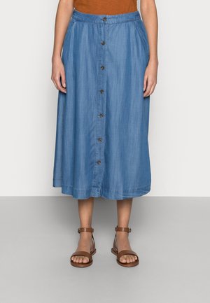 Jupe midi bleue avec boutons sur le devant, poches, portée avec des sandales marron à bride de cheville sur pieds nus.