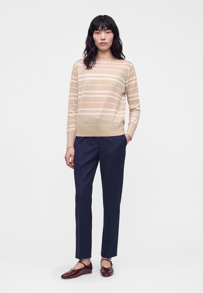 Femme debout les mains dans les poches, vêtue d'un pull à manches longues beige et blanc rayé, d'un pantalon bleu marine et de chaussures Mary Jane bordeaux.